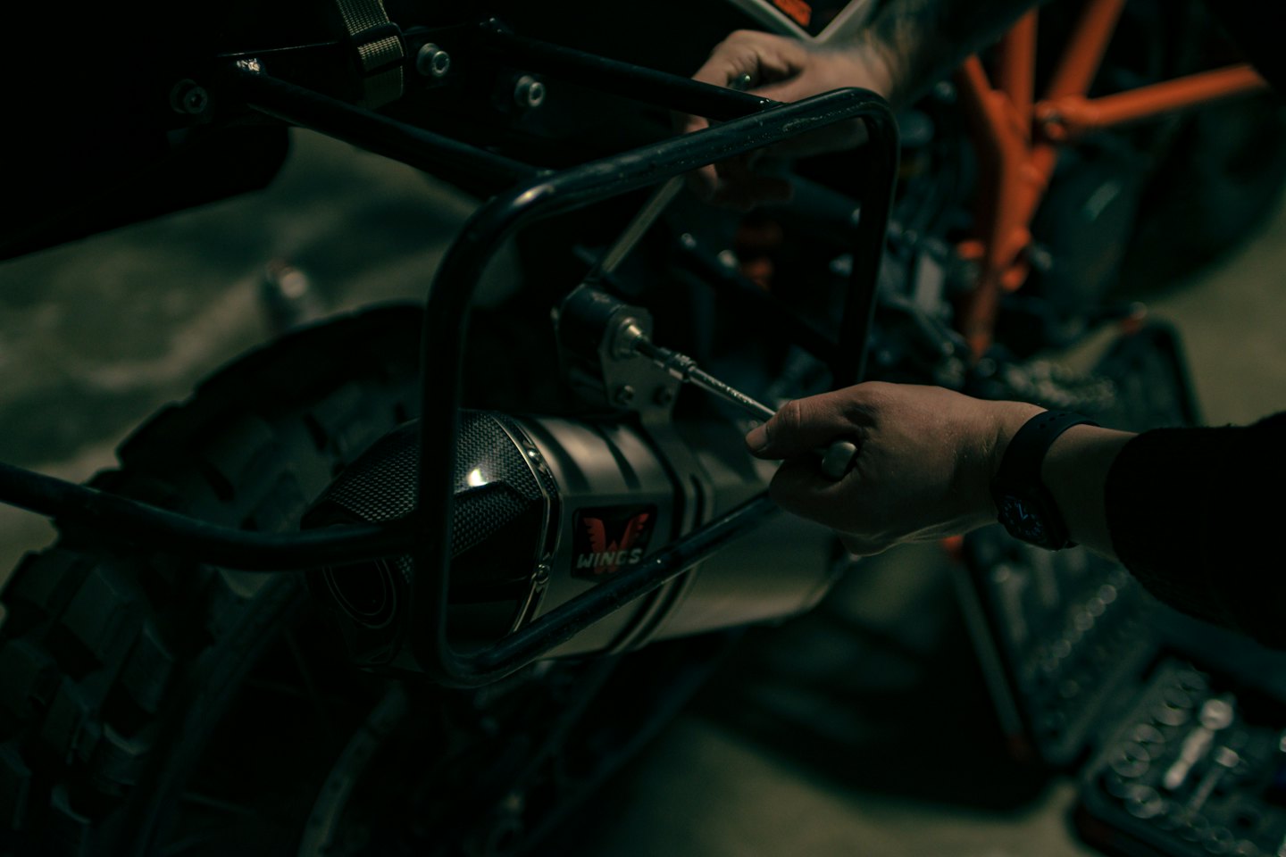 Mechanic's hands repair a motorcycle exhaust with a wrench.